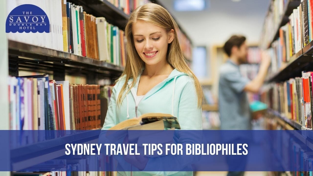 A smiling young woman in a light green hoodie reading an open book in a library or bookstore aisle.