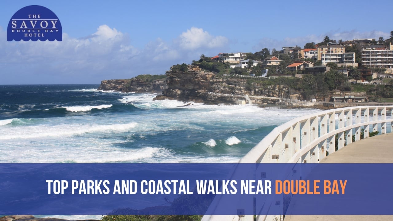 A sunny, high-angle view of ocean waves crashing against a rugged, rocky cliffside with residential buildings along the ridge.