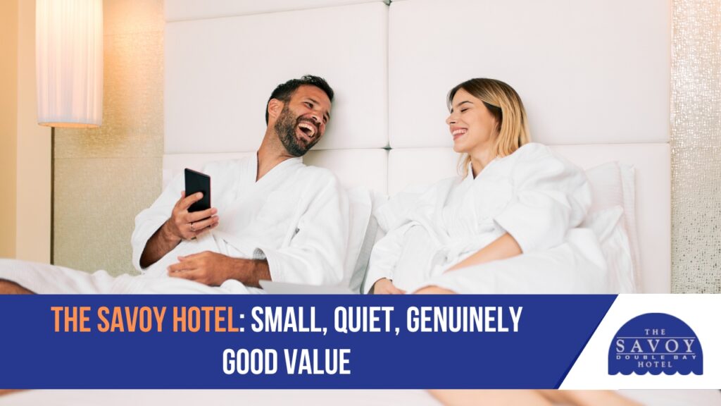 Experience the Art of Slow Travel at Double Bay Hotel - Double Bay A couple in white bathrobes relaxes and laughs in a hotel bed while looking at a smartphone.